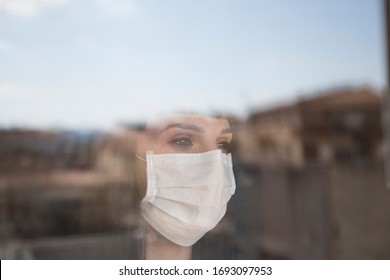 Brunette Woman With Mask Looking Out The Window During Home Confinement. With The City Reflected In The Glass. Looking Forward To Going Outside. Concept Covid 19.