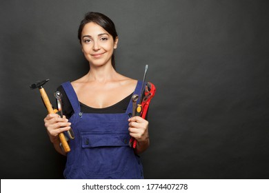 Brunette Woman With Mechanic Work Tools In Hand And Blue Overalls Makes Face Amused, Isoalta On Black Background
