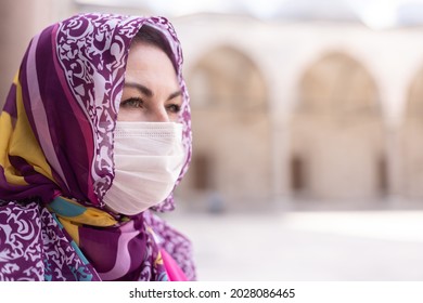 Brunette woman in a protective mask on her face and a scarf on her head against the background of a building with arches