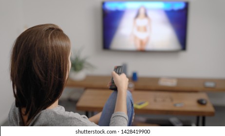 Brunette Woman Is Resting On A Couch In Home And Watching Tv Fashion Show, Holding Remote Control In Hand