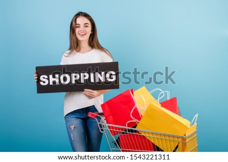 brunette woman with shopping cart full of colorful red and yellow paper bags and shopping sign isolated over blue
