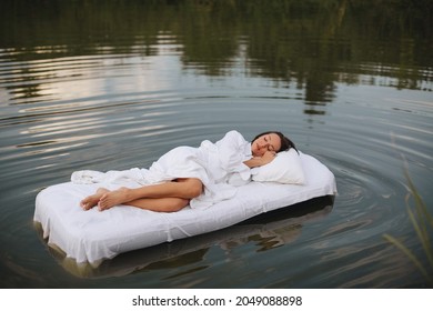 Brunette woman with short hair in a white shirt in bed on the water. A young woman floats on an inflatable mattress with a white sheet on the lake.