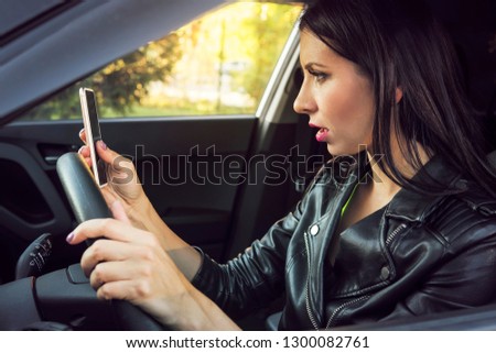 Brunette woman sitting inside car with mobile phone in hand texting while driving. Shocked girl checking her smartphone not paying attention at road stunned by bad text message. Dangerous distraction