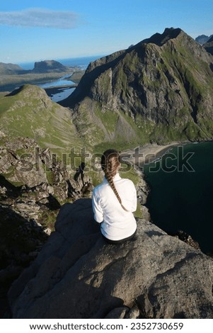 Brunette woman sitting on the cliff at the top of Ryten mountain in Lofoten islands, Northern Norway. She is wearing a white sports shirt and black shorts. Braided hair. Blue sky, carefree hiking