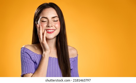 Brunette Woman Smiling From Pleasure. Girl With Closed Eyes, Touches Her Face And Dreams About Smth, Thinking Or Imaging, Standing Over Orange Background