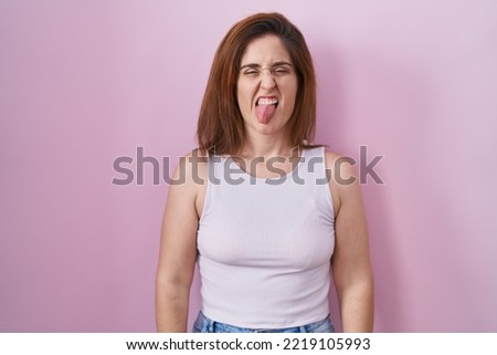 Brunette woman standing over pink background sticking tongue out happy with funny expression. emotion concept. 