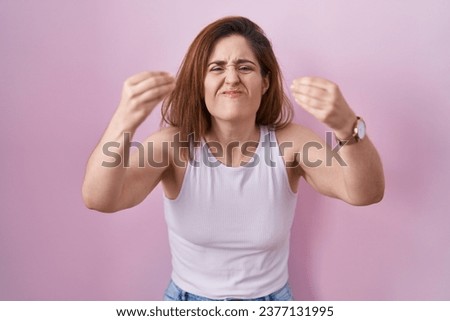 Brunette woman standing over pink background angry and mad raising fists frustrated and furious while shouting with anger. rage and aggressive concept. 