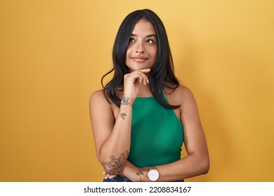 Brunette Woman Standing Over Yellow Background With Hand On Chin Thinking About Question, Pensive Expression. Smiling And Thoughtful Face. Doubt Concept. 