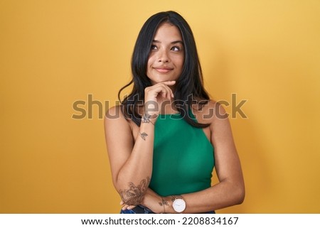 Brunette woman standing over yellow background with hand on chin thinking about question, pensive expression. smiling and thoughtful face. doubt concept. 