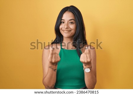 Brunette woman standing over yellow background doing money gesture with hands, asking for salary payment, millionaire business 