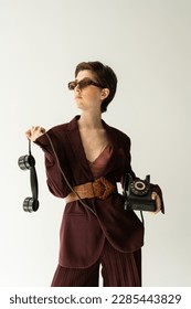 brunette woman in sunglasses and burgundy brown jacket with leather belt posing with vintage telephone isolated on grey - Shutterstock ID 2285443829