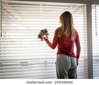 A Brunette Woman In Sweat Pants Using A Feather Duster On White Blinds 