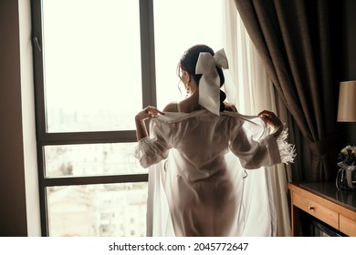 A Brunette Woman Takes Off Her White Transparent Robe In A Hotel Room Against The Background Of A Window. Young Sexy Woman With White Ribbon In Her Hair Undresses And Looks Out The Window