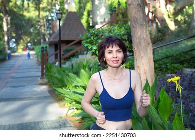 A brunette woman in a tracksuit runs in the morning along a path among palm trees and pines. - Shutterstock ID 2251430703