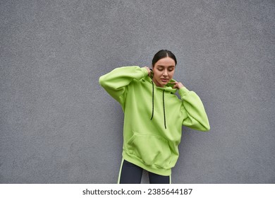 brunette woman in trendy lime color hoodie adjusting hood and standing near grey concrete wall - Powered by Shutterstock