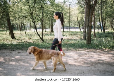 Brunette Woman Walking On Park Alley With Dog. Side View Of Girl In White Blouse Walking Her Dog In Park During Sunny Day.