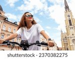 Brunette woman wearing sunglasses riding a bike, bicycle travel tour around historic place in Novi Sad, Serbia.