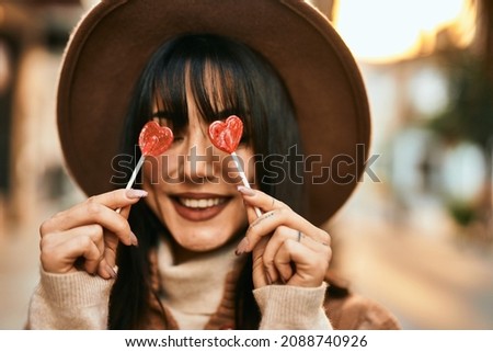 Brunette woman wearing winter hat being funny holding lollipops covering eyes outdoors at the city