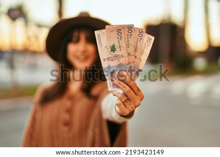 Brunette woman wearing winter hat smiling holding colombian pesos banknotes outdoors at the city on sunset