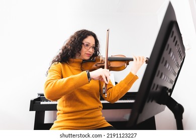 Brunette Woman Wearing A Yellow Sweater, And Playing Violin By Reading Sheet Music.