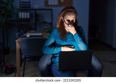 Brunette Woman Working At The Office At Night Smelling Something Stinky And Disgusting, Intolerable Smell, Holding Breath With Fingers On Nose. Bad Smell 