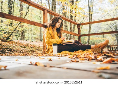  Brunette Frau in gelbem Pullover, die auf einem gefallenen Herbst sitzt, hinterlässt in einem Park ein Buch oder schreibt ein Tagebuch
