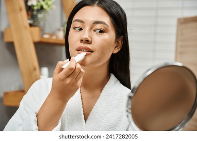 brunette young asian woman in bath robe applying lip balm and looking at mirror in bathroom - Powered by Shutterstock