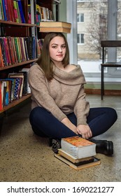 Brunette young girl sitting on the floor and balancing stack of books on her head near bookshelf in library. Plus size model