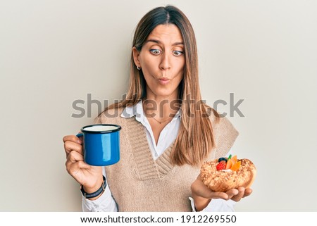 Brunette young woman drinking coffee and eating pastry making fish face with mouth and squinting eyes, crazy and comical. 