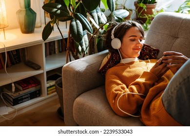 Brunette young woman listening music and using cellphone while resting on couch at home