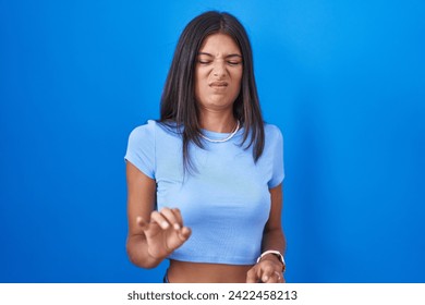 Brunette young woman standing over blue background disgusted expression, displeased and fearful doing disgust face because aversion reaction. 