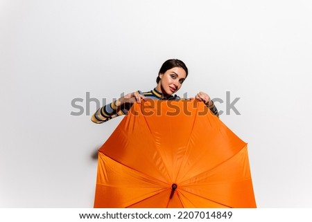 brunette young woman in striped turtleneck holding orange umbrella on grey