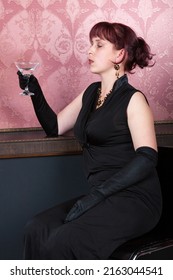 brunette young woman wearing black gown and long black gloves is sitting on a bar chair with glass of vodka in her hand