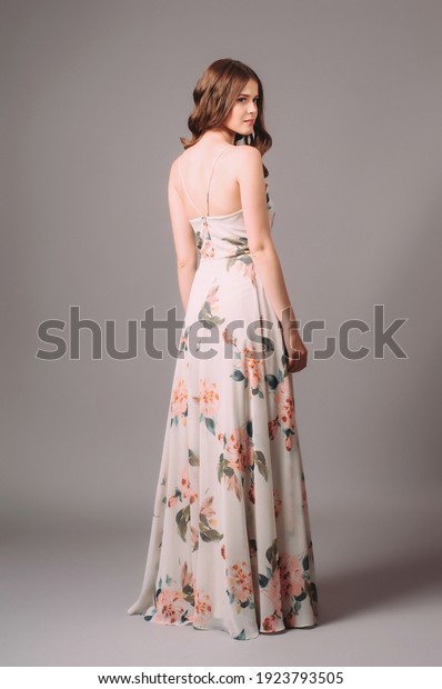 Brunette young woman in white sundress with\
floral print. Calm studio portrait of young lady in long backless\
evening gown on grey\
background.