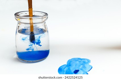 A brush with blue paint is washed in a glass jar with water, blue cloud. Paint dripping into water, white sheet of paper, blue cloud painted with watercolors on canvas, space for copy - Powered by Shutterstock