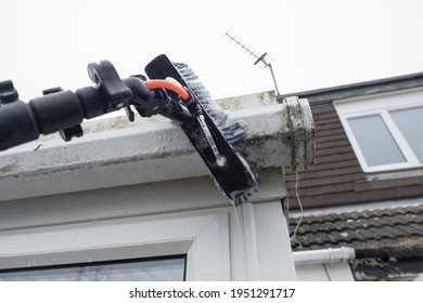 A brush cleaning dirty clogged white plastic pvc gutters and drain pipes with mossy green mould plastic fascias.  Blocked drains and guttering need window cleaners and regular yard work maintenance  - Shutterstock ID 1951291717