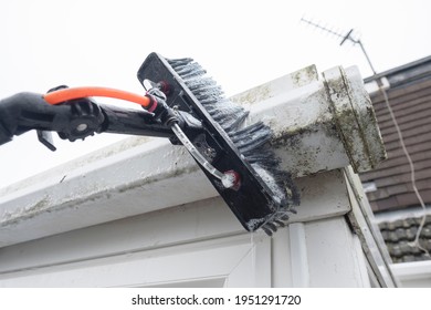 A brush cleaning dirty clogged white plastic pvc gutters and drain pipes with mossy green mould plastic fascias.  Blocked drains and guttering need window cleaners and regular yard work maintenance 