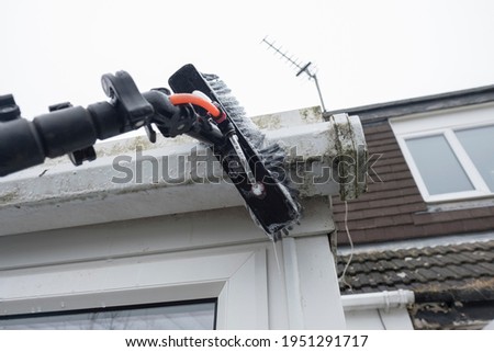 A brush cleaning dirty clogged white plastic pvc gutters and drain pipes with mossy green mould plastic fascias.  Blocked drains and guttering need window cleaners and regular yard work maintenance 