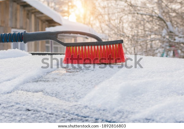 Brush for cleaning snow from a car close-up. A brush\
with red bristles against the backdrop of sunlight stands on a\
snow-covered car. Concept - cleaning the car from snow, frost,\
blizzard, cold