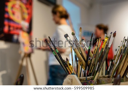 Brushes for painting. Various shapes of bristle, differents handlers of artistic tools. Unfocused background with students working in the art studio.