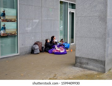 Brussels, Belgium November 14, 2019: 
Poverty: Refugees, Slavic Type Sleeping Baby On Mother's Womb, Teen Daughter Sitting Against A Financial Government Building Granite Wall Begging For Some Money.