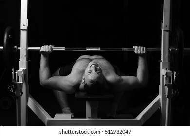 Brutal Athletic Man Pumping Up Muscles On Bench Press In Monochrome. Bodybuilder. Naked To The Waist. Black And White Photography. Sport. Low Key. Gym. Isolated On Black.