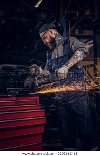 Brutal auto mechanic in a uniform and safety\
glasses working with an angle grinder while standing against a\
broken car in repair\
garage.
