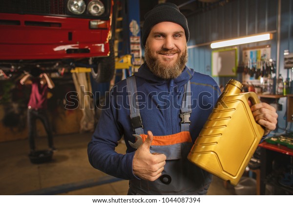 Brutal bearded car mechanic works in the garage. The\
master advises the engine oil. Happy mechanic car with bottle of\
gold engine oil.