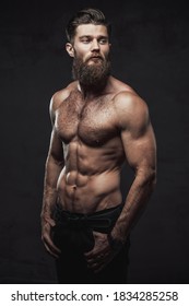 Brutal bearded guy with naked torso and muscular build with hands in pockets posing in dark studio background.