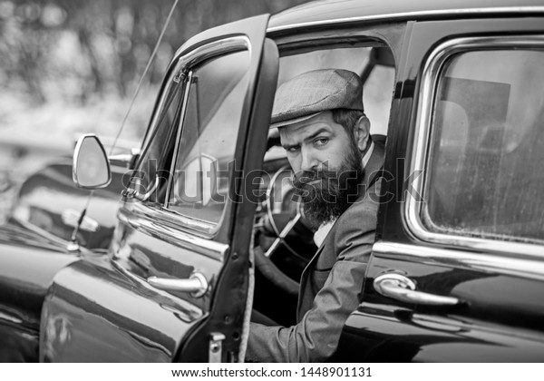 brutal bearded man with a mustache in a cap in the\
retro car looking at\
camera