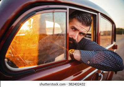 Brutal Bearded Man With A Mustache In A Shirt, Pants With Suspenders With A Girl With Dark Hair And Big Lips With Bright Red Lipstick In A Short Dress And Heels Near Retro Car At Sunset