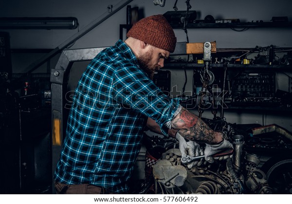 A brutal bearded man with tattooed arm\
repairing the car\'s engine in a\
garage.