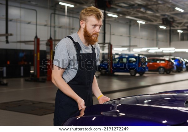 A brutal bearded\
man in uniform lifts the hood of a car to carry out repair work.\
Modern diagnostic station