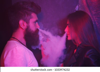 Brutal bearded man and young pretty woman blowing smoke to join it in one cloud. Relationship and vape addiction concept. Couple smoke an electric cigarette on dark and red light room. Couple vaping